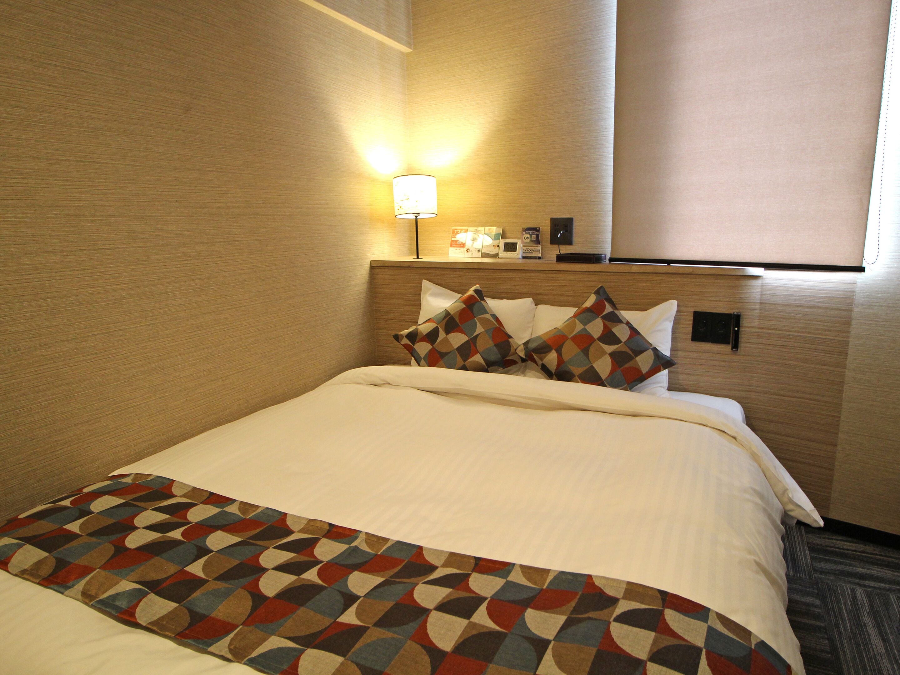 A hotel room with a neatly made bed, a patterned throw blanket, and a bedside lamp.