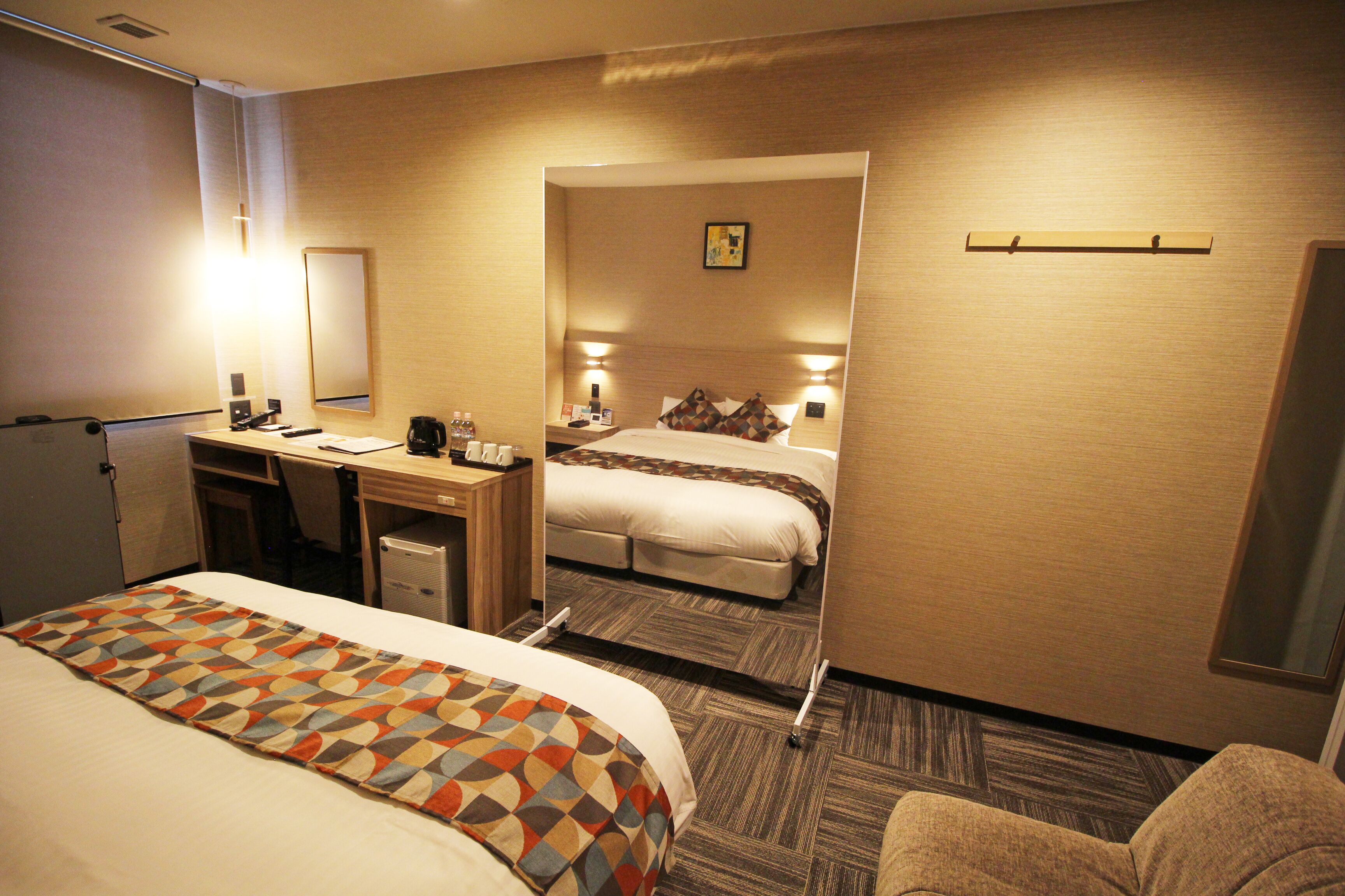 A hotel room with a large bed, a desk with a chair, a small table with a lamp, and a mirror on the wall.