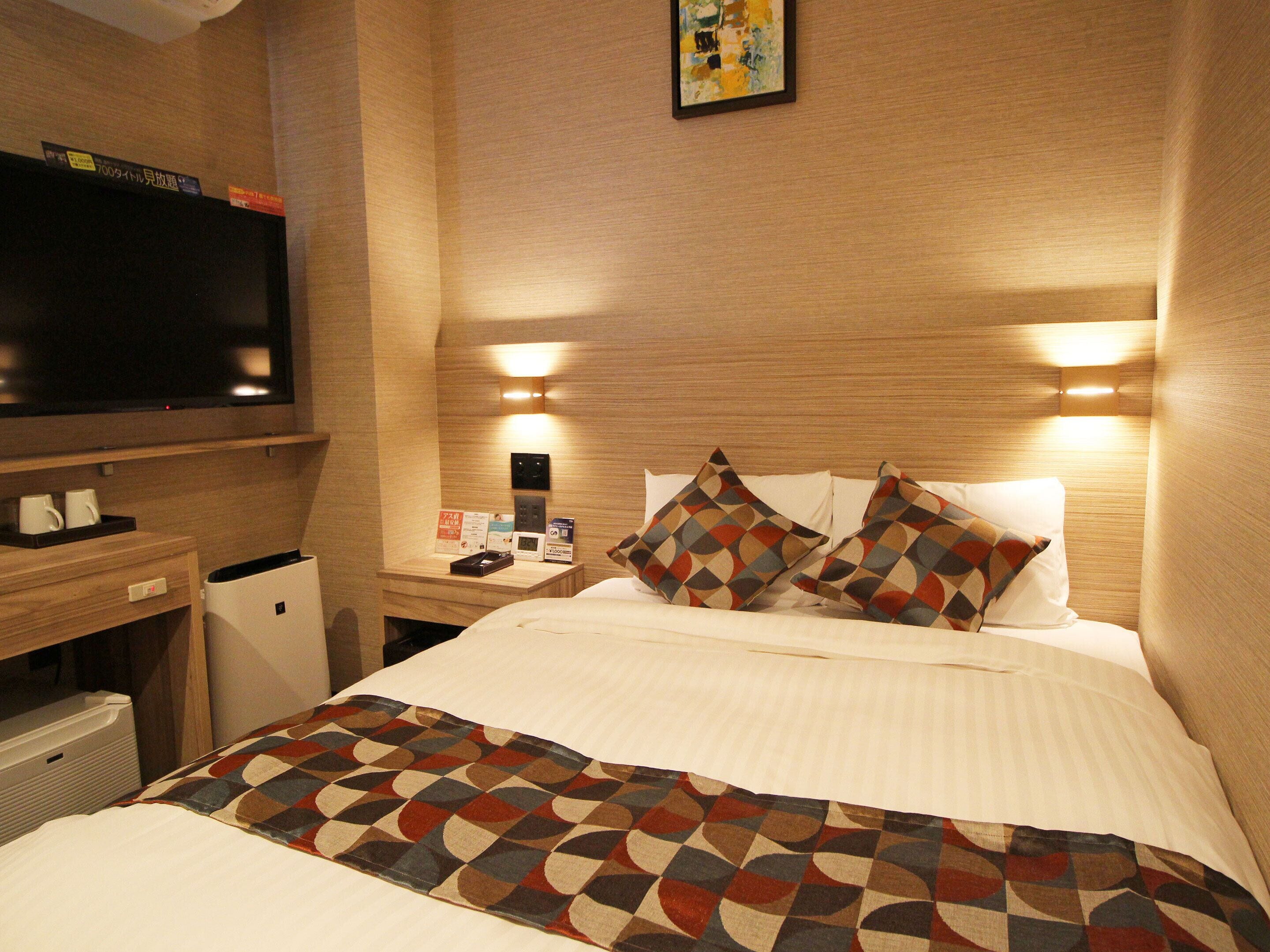 A hotel room with a large bed, a flat-screen TV, a desk with a computer, and a nightstand with a lamp and a clock.