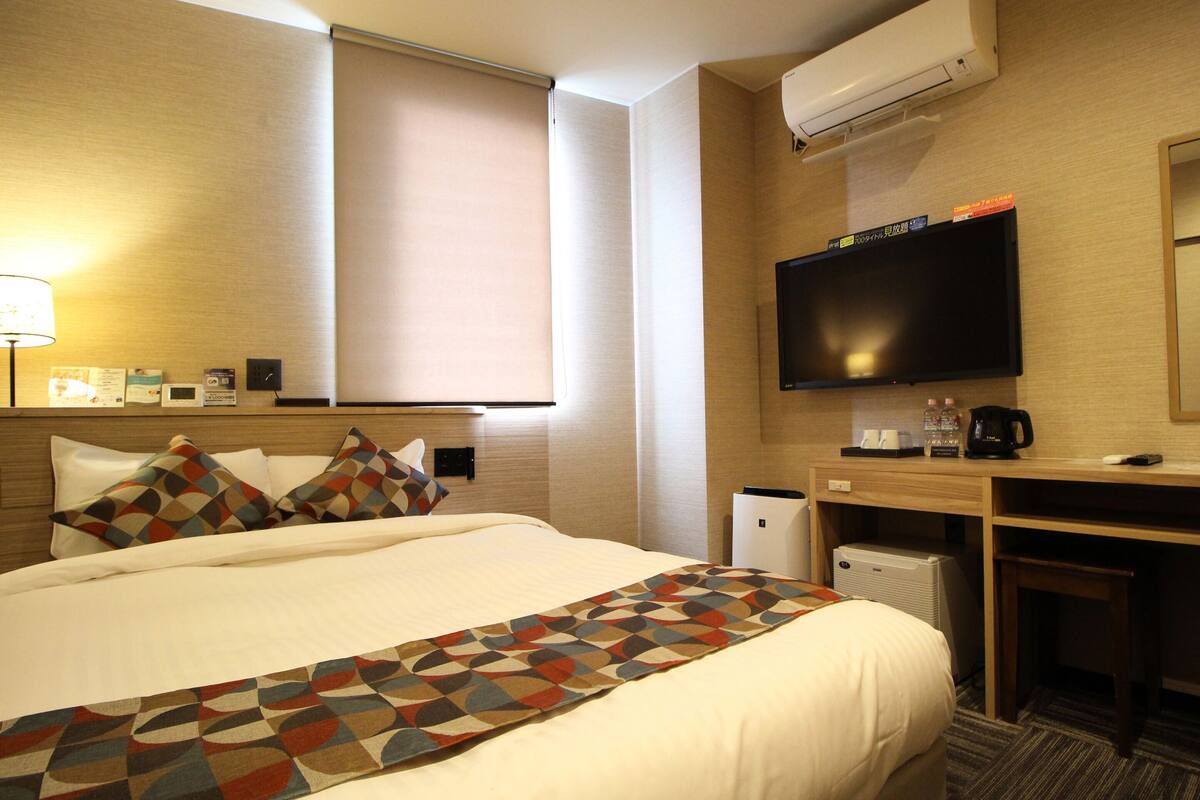 A hotel room with a bed, a desk, a television, and an air conditioning unit.