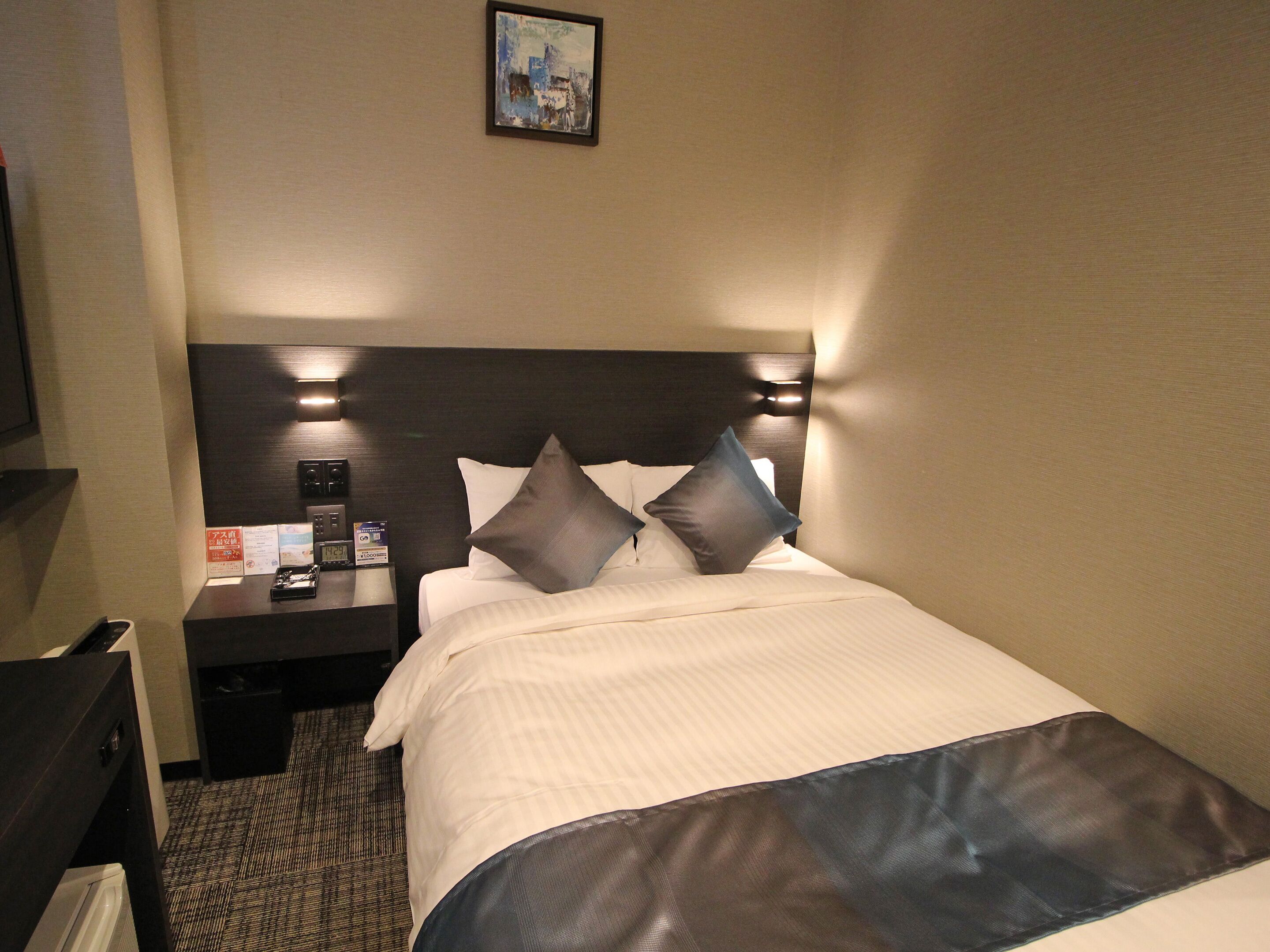 Une chambre d’hôtel avec un lit bien fait, une table de chevet avec une horloge et un téléphone, et une lampe fixée au mur.