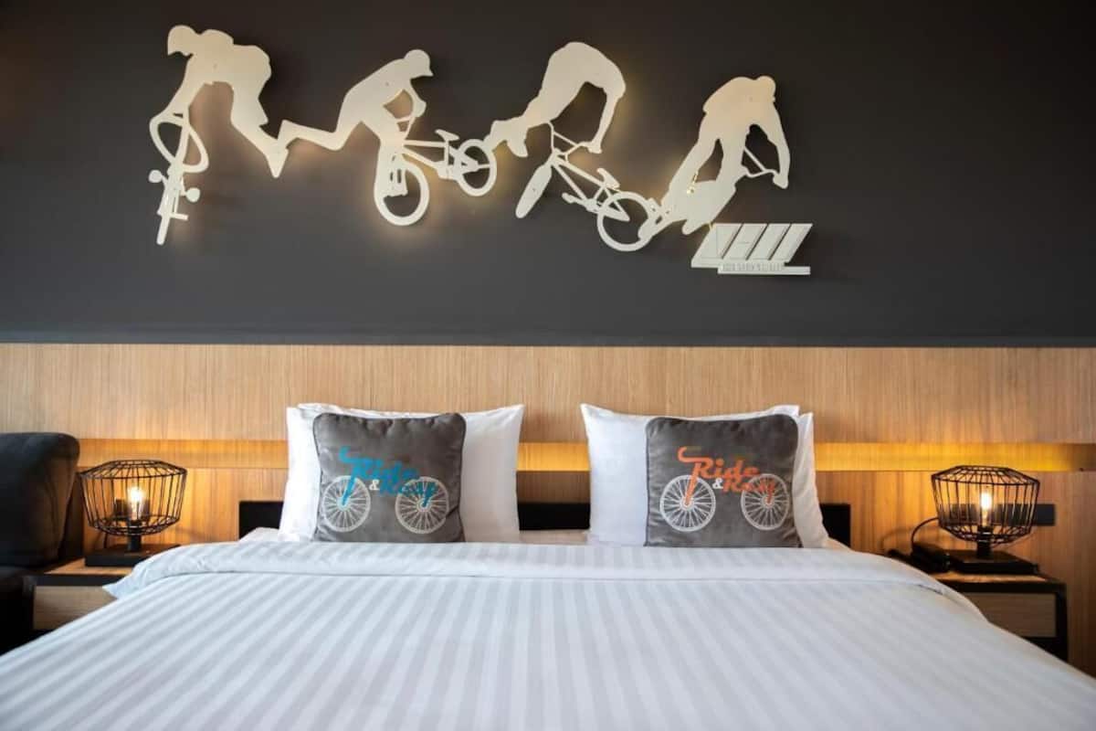 A hotel room with a bed, two nightstands, a wall-mounted light fixture, and a wall art piece depicting cyclists.