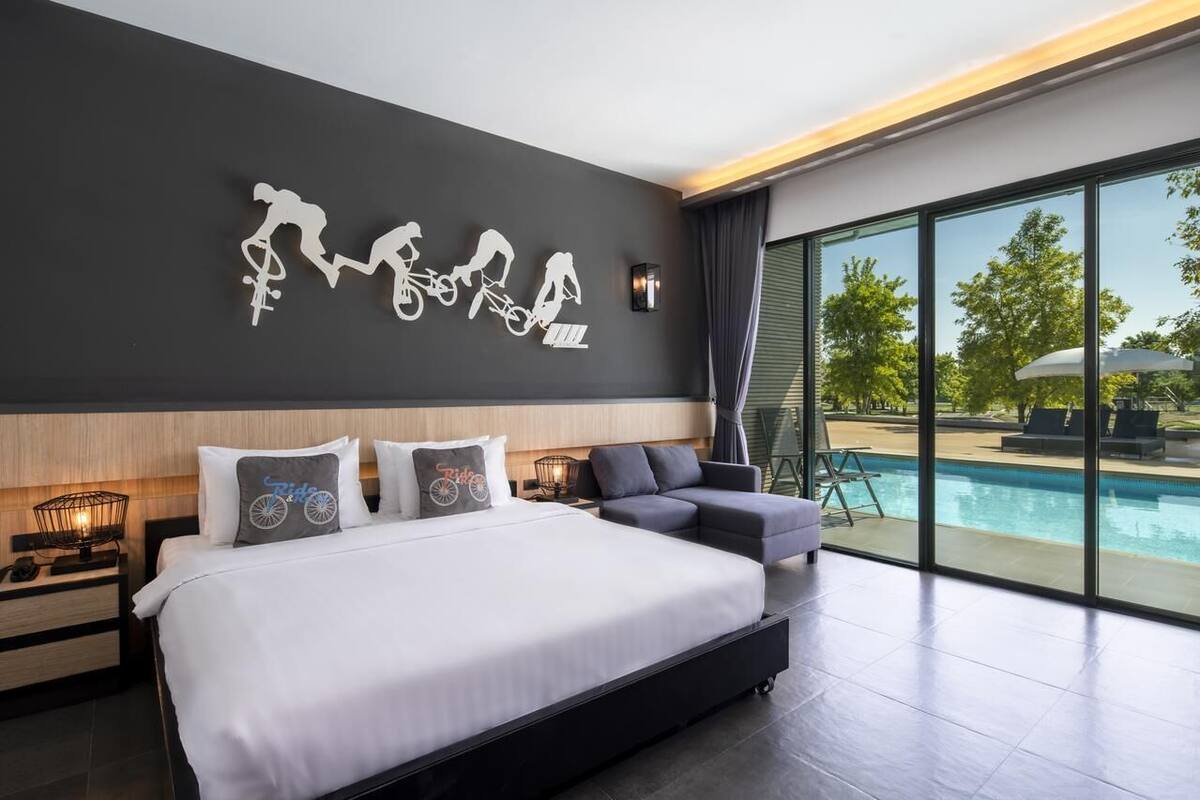 A modern hotel room with a large bed, a wall-mounted bike art piece, a view of an outdoor swimming pool, and a balcony with outdoor furniture.