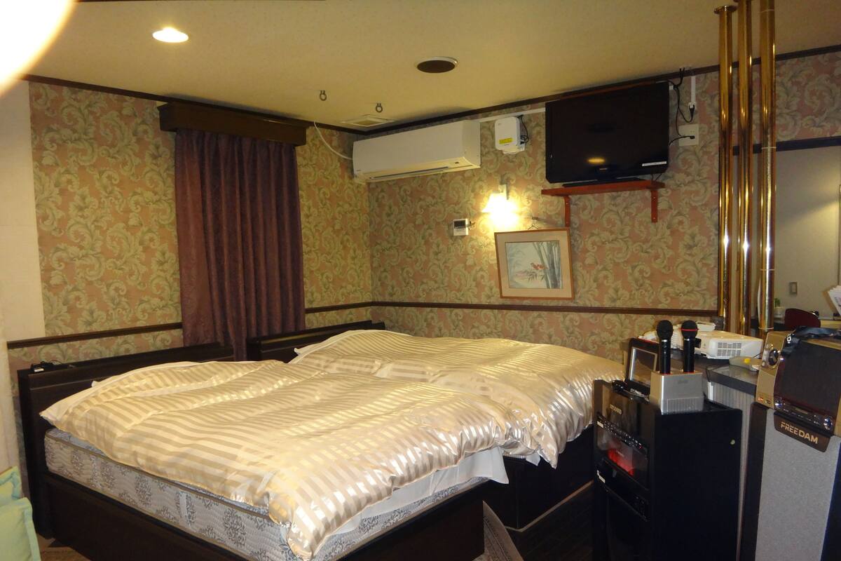 Habitació d'hotel amb un llit gran, televisió penjada a la paret, aparell d'aire condicionat i escriptori amb micròfon i altre equipament.