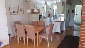 A modern kitchen with white cabinets, a wooden dining table with four chairs, pendant lights, and a built-in oven. - Kotimaailma Apartments Naantali (Naantali)