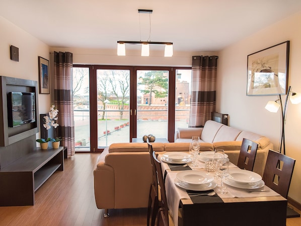 A modern living room with a sofa, dining table set for four, and a flat-screen TV mounted on the wall. - VacationClub Apartments - Morska (Kolobrzeg)