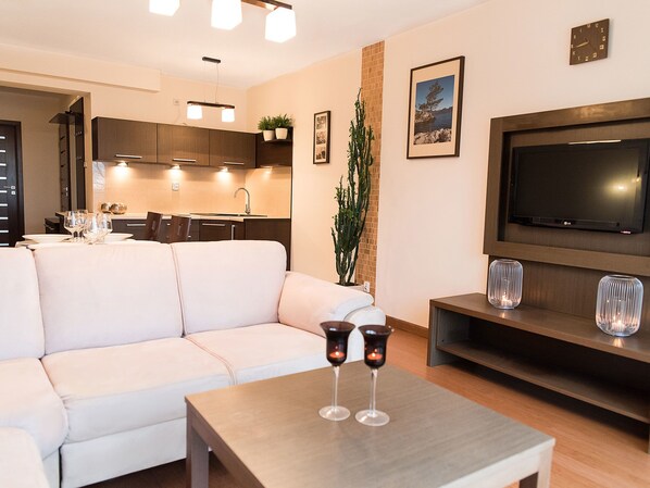 A modern living room with a sofa, a coffee table with glasses, a TV, and a kitchen in the background.