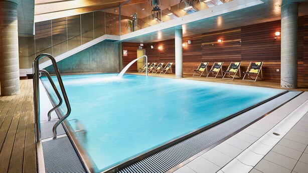 An indoor swimming pool with a clear water surface, a metal ladder, and a tiled floor.