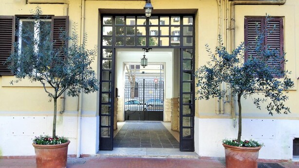 Entrada de un edificio con una verja negra, dos plantas en macetas a cada lado y un camino empedrado que conduce al interior.