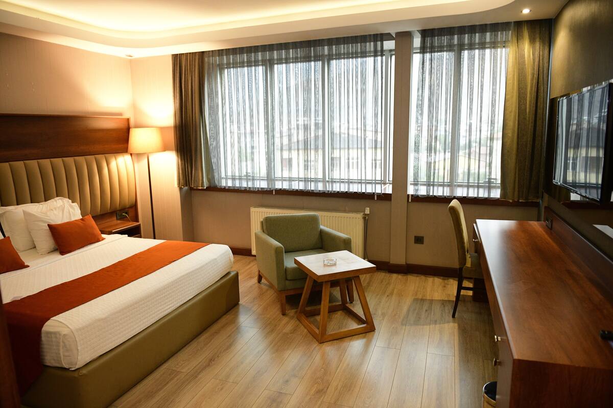 Une chambre d’hôtel avec un grand lit, un bureau, une chaise et une vue sur l’extérieur à travers de fines rideaux.