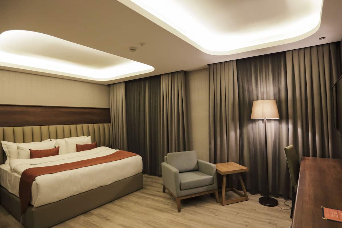 Une chambre d’hôtel avec un grand lit, un petit fauteuil, une table de chevet en bois, une lampe et un téléviseur à écran plat fixé au mur.
