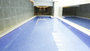 Une grande piscine intérieure, dont le sol et les murs sont recouverts de carreaux bleus, et qui est entourée de murs en pierre grise.