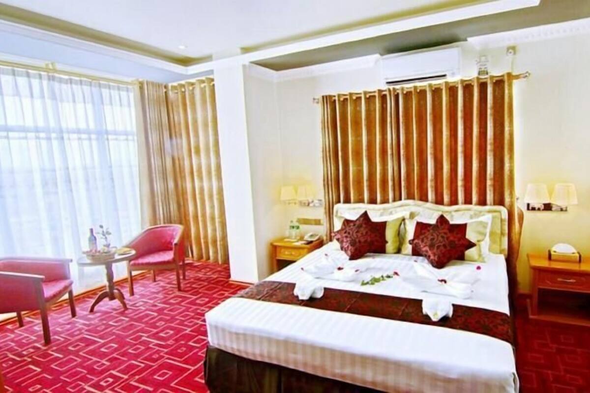 A hotel room with a large bed, two bedside tables with lamps, a small table with a chair, and a window with curtains.
