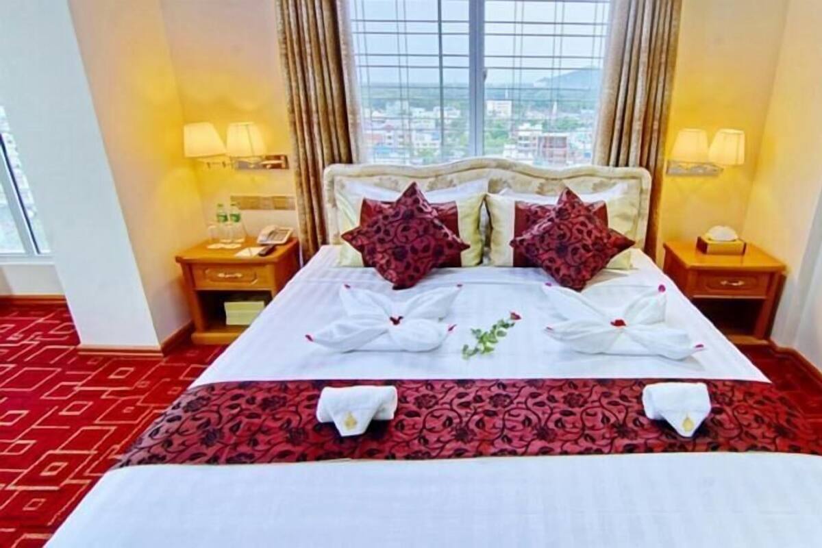 A hotel room with a large bed, two nightstands, a window with a city view, and a red patterned carpet.