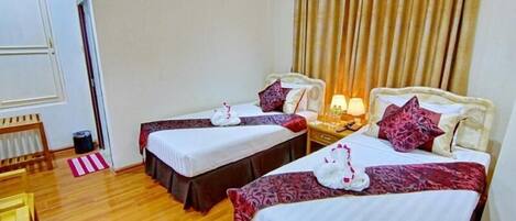 Habitación de hotel con dos camas, cada una con una colcha blanca y una almohada decorativa roja. El suelo es de madera, hay una mesita con una lámpara y una ventana con cortinas.
