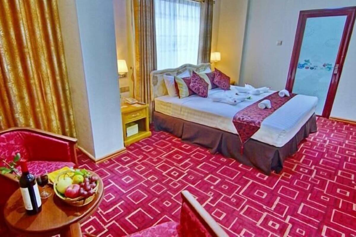 A hotel room with a bed, a red armchair, a small table with a fruit basket, and a patterned carpet.