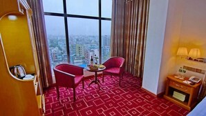 Quarto de hotel com uma grande janela, duas cadeiras vermelhas, uma mesa pequena com uma garrafa e frutas, e uma mesa de cabeceira com um abajur e um telefone.