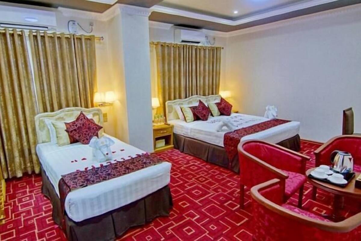 A hotel room with two beds, a red patterned carpet, a red armchair, and a small table with a kettle and cups.