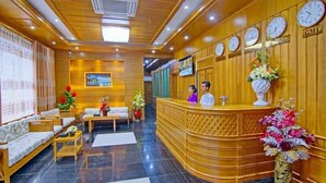 Um hall de hotel com balcão de recepção, painéis de madeira, relógios e área de estar com sofás e mesas.