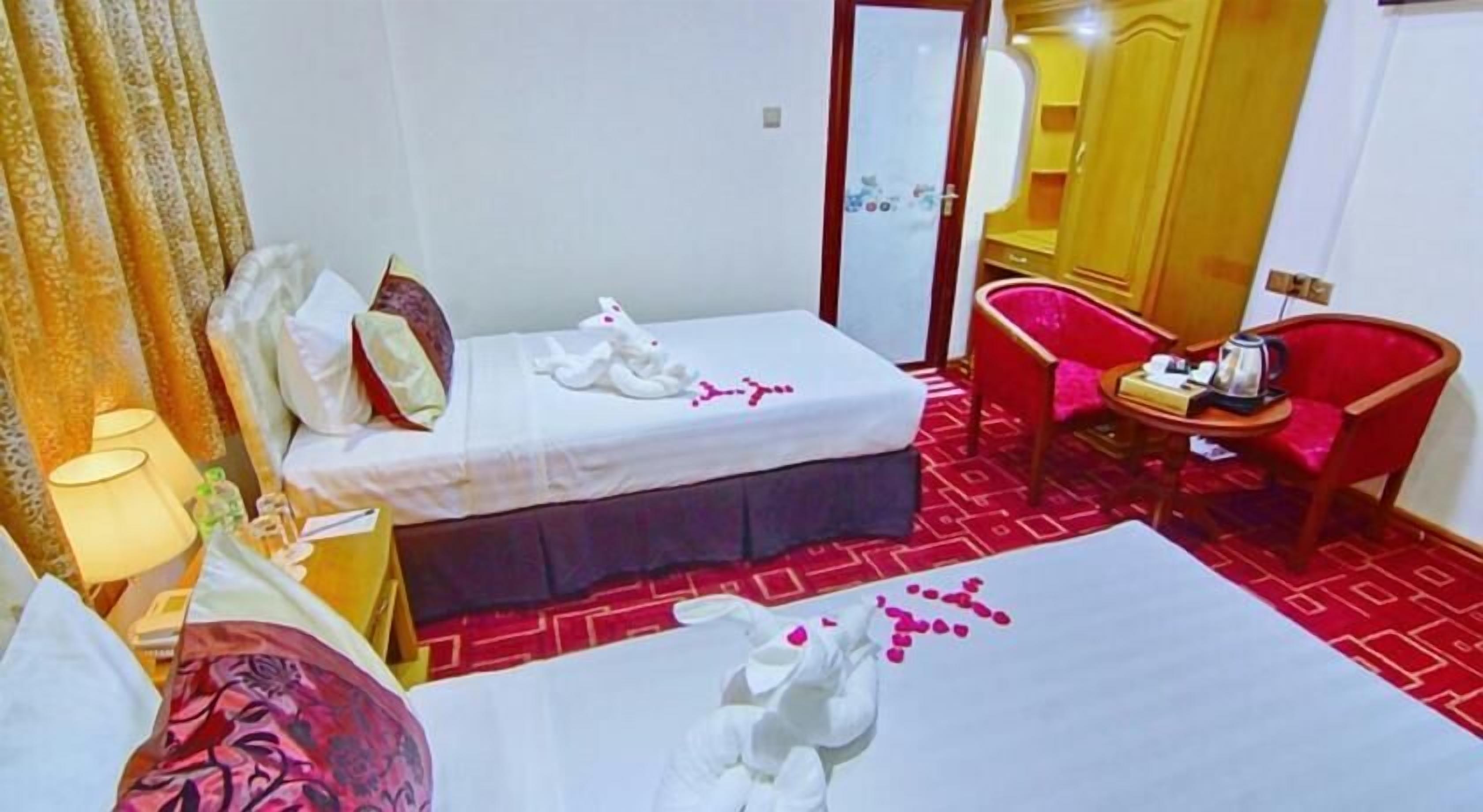 Een hotelkamer met twee bedden, een rood tapijt, een rode stoel, een klein tafeltje met een waterkoker en een lamp.