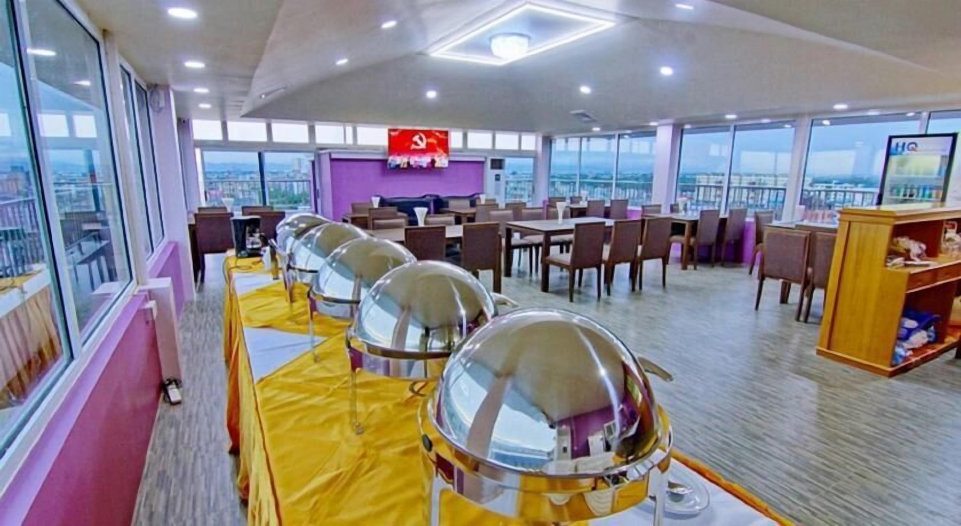 A dining area with round tables, a buffet setup, and a large window offering a city view.