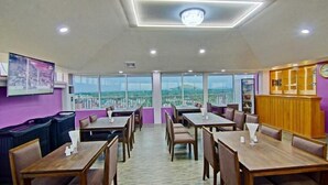 Uma área de refeições com mesas e cadeiras, grandes janelas com vista para a cidade e um relógio na parede.