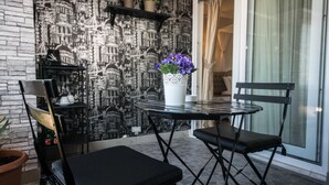 Un pequeño espacio al aire libre con una mesa redonda y dos sillas, una planta en maceta y una pared decorativa con motivos de paisaje urbano.