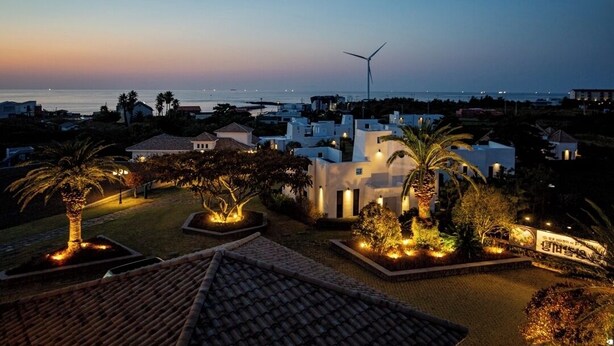 A coastal residential area at dusk with houses, a wind turbine, and well-lit gardens.