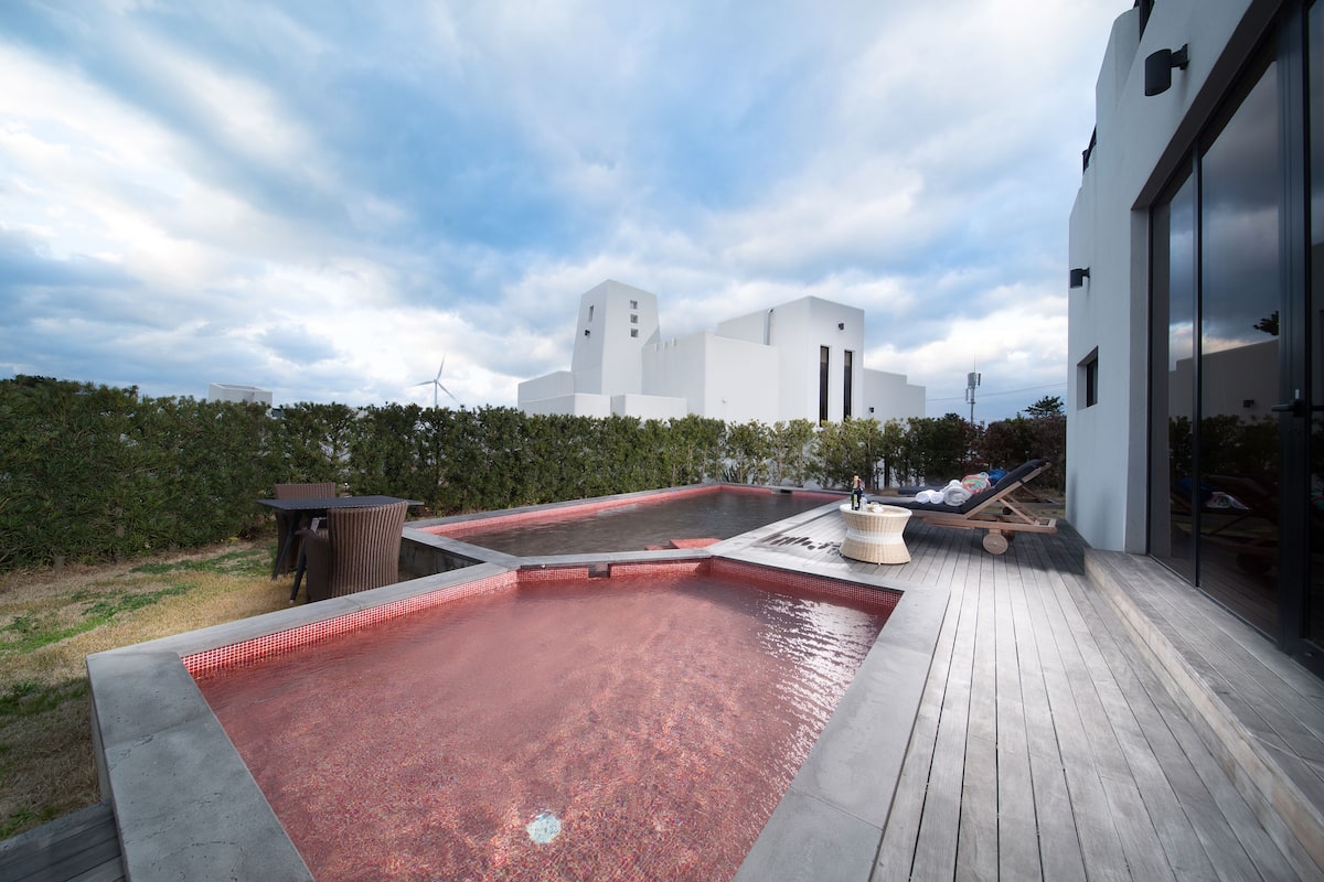 A rooftop pool with a wooden deck, lounge chairs, and a view of a white building with a tower.
