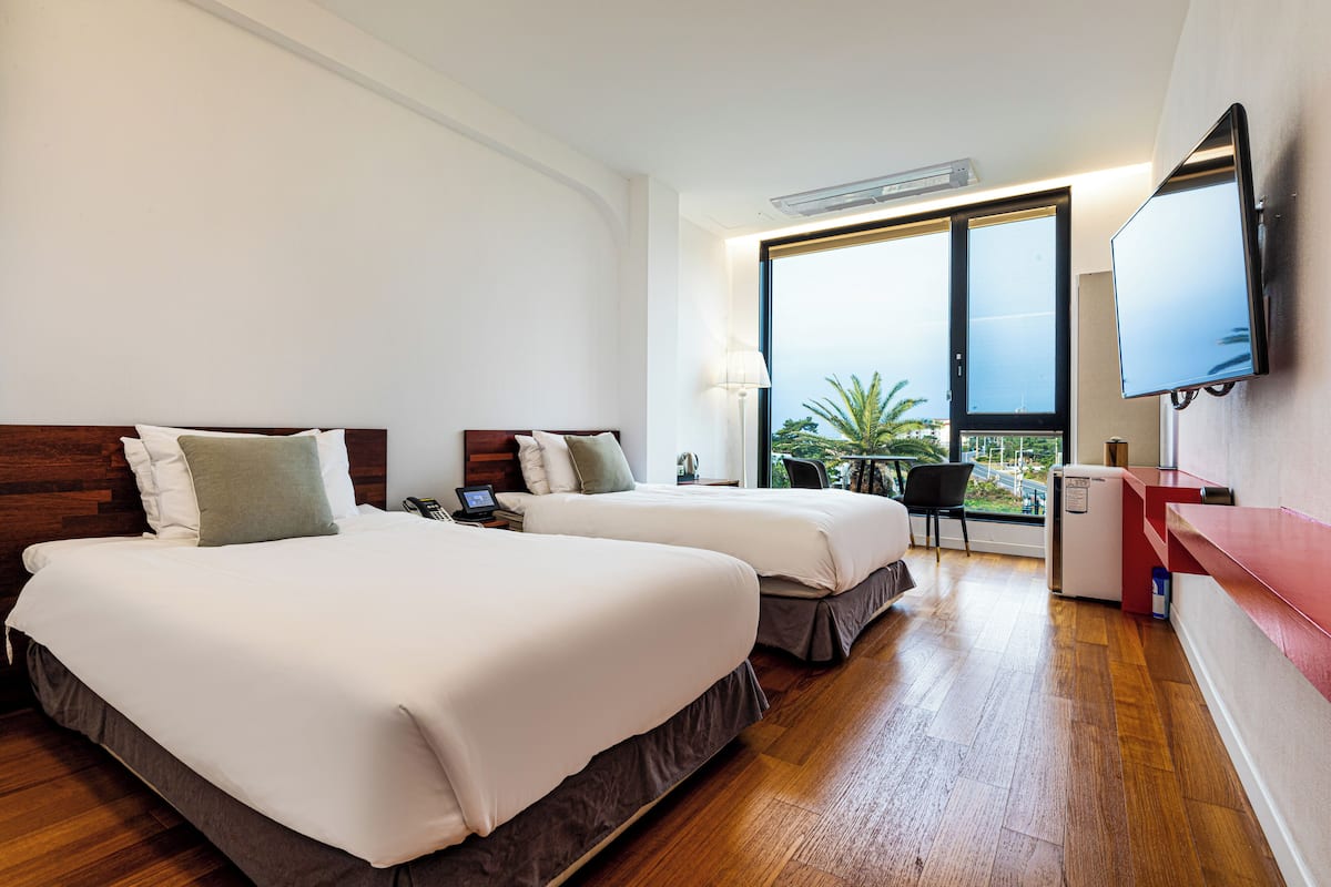 A hotel room with two beds, a desk, a chair, a TV, and a window with a view of palm trees.