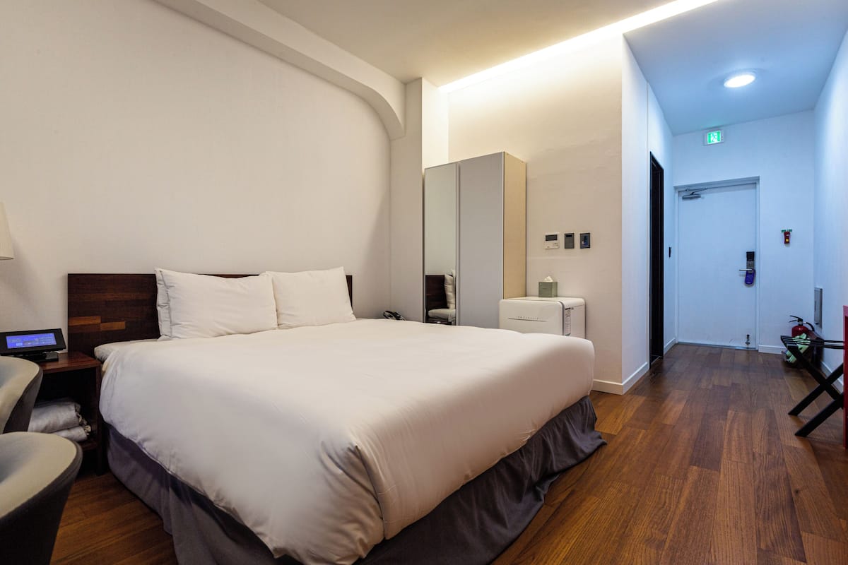 A hotel room with a large bed, a desk with a computer, a chair, a wardrobe, and a bathroom with a toilet and sink.
