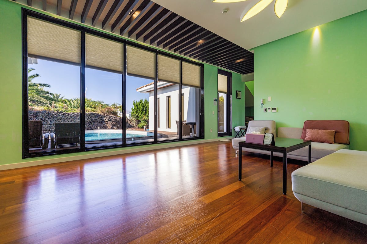 A spacious living area with a wooden floor, a large sliding glass door leading to a pool, and a green wall.