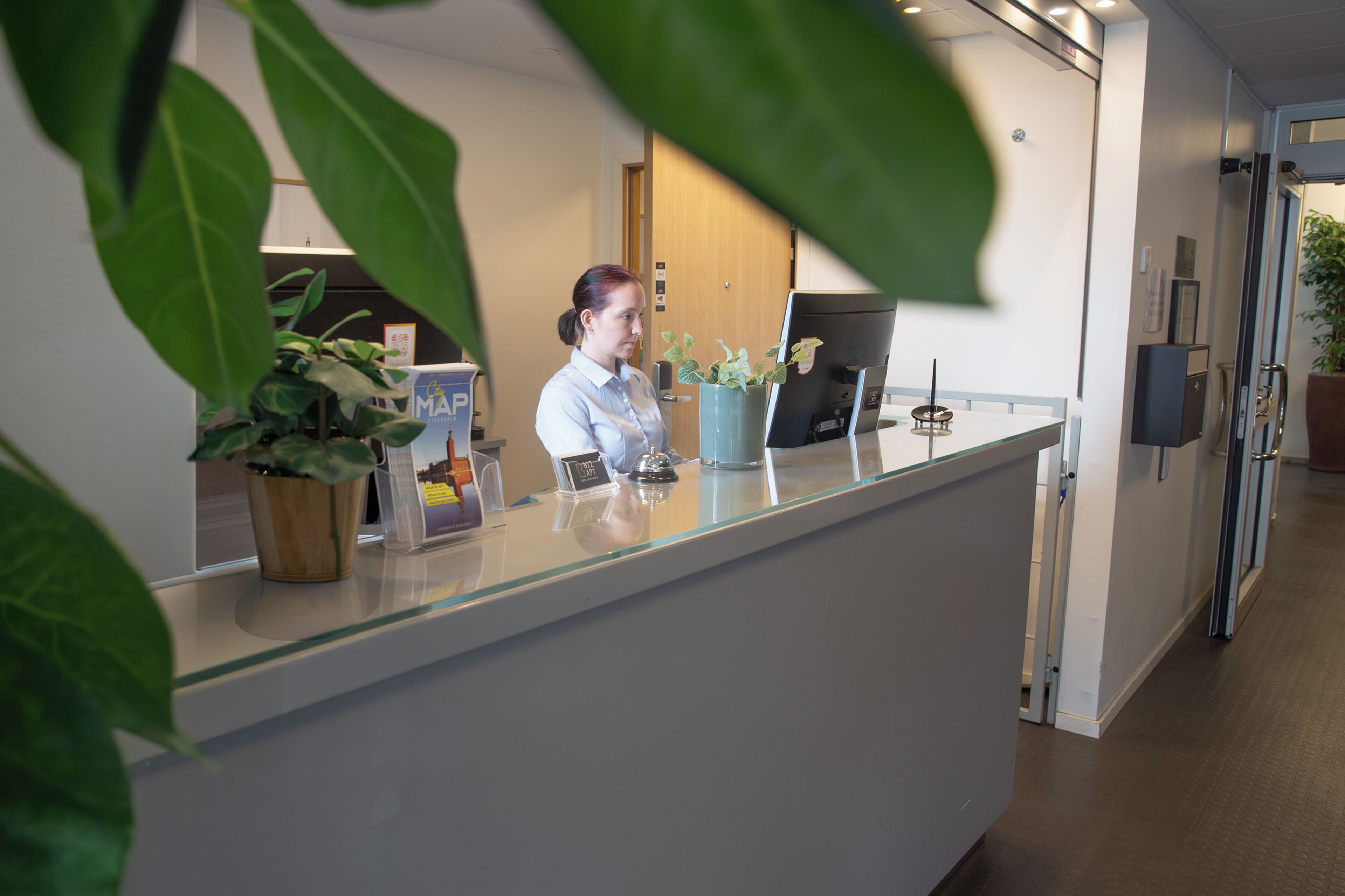 A reception desk with a receptionist, a potted plant, a computer, and a glass partition.