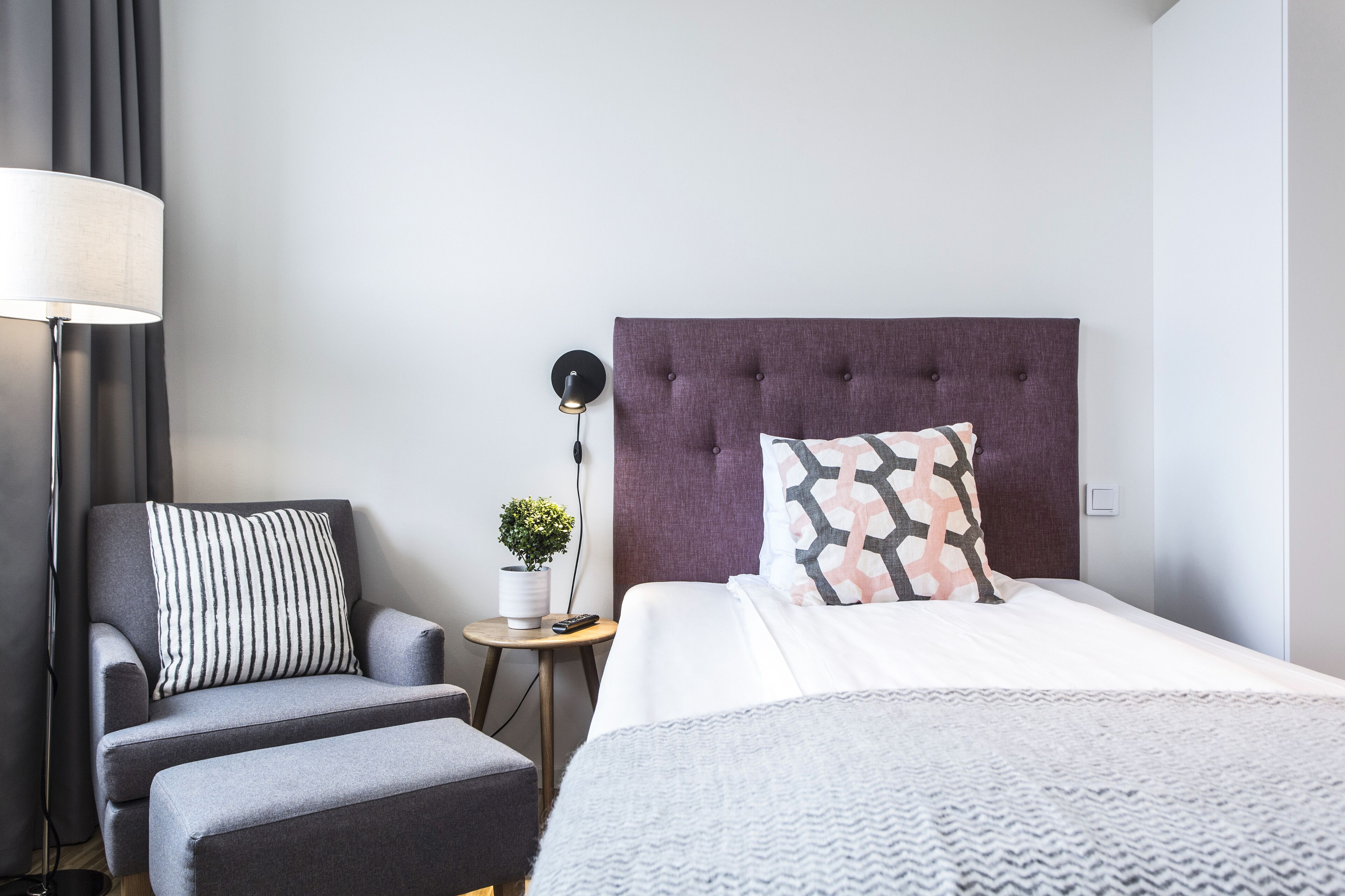 Ett modernt hotellrum med en grå soffa, ett litet runt bord med en lampa, en krukväxt och en säng med en vit och mönstrad kudde.
