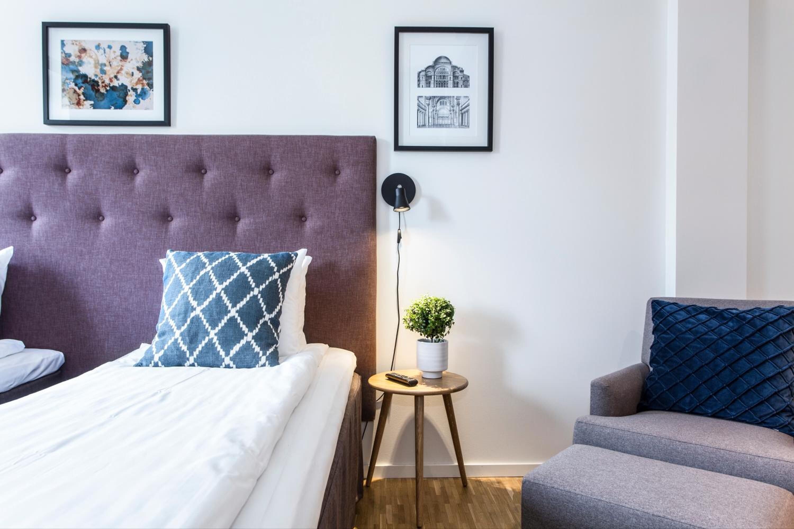 A bedroom with a tufted headboard, a bed with white and blue patterned pillows, a small round table with a plant, and a chair with a blue pillow.