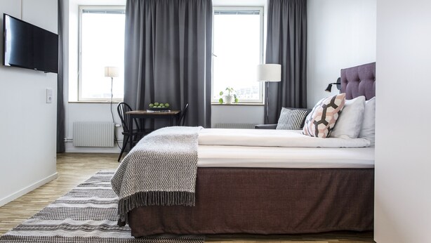 Une chambre d’hôtel moderne dotée d’un grand lit, d’un téléviseur à écran plat fixé au mur, d’un bureau avec une lampe et offrant une vue sur la ville grâce à de grandes fenêtres.