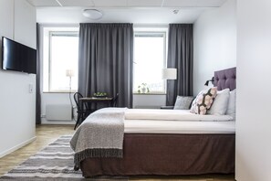 A modern hotel room with a large bed, a flat-screen TV mounted on the wall, a desk with a lamp, and a view of the city through large windows. - Biz Apartment Solna (Solna)