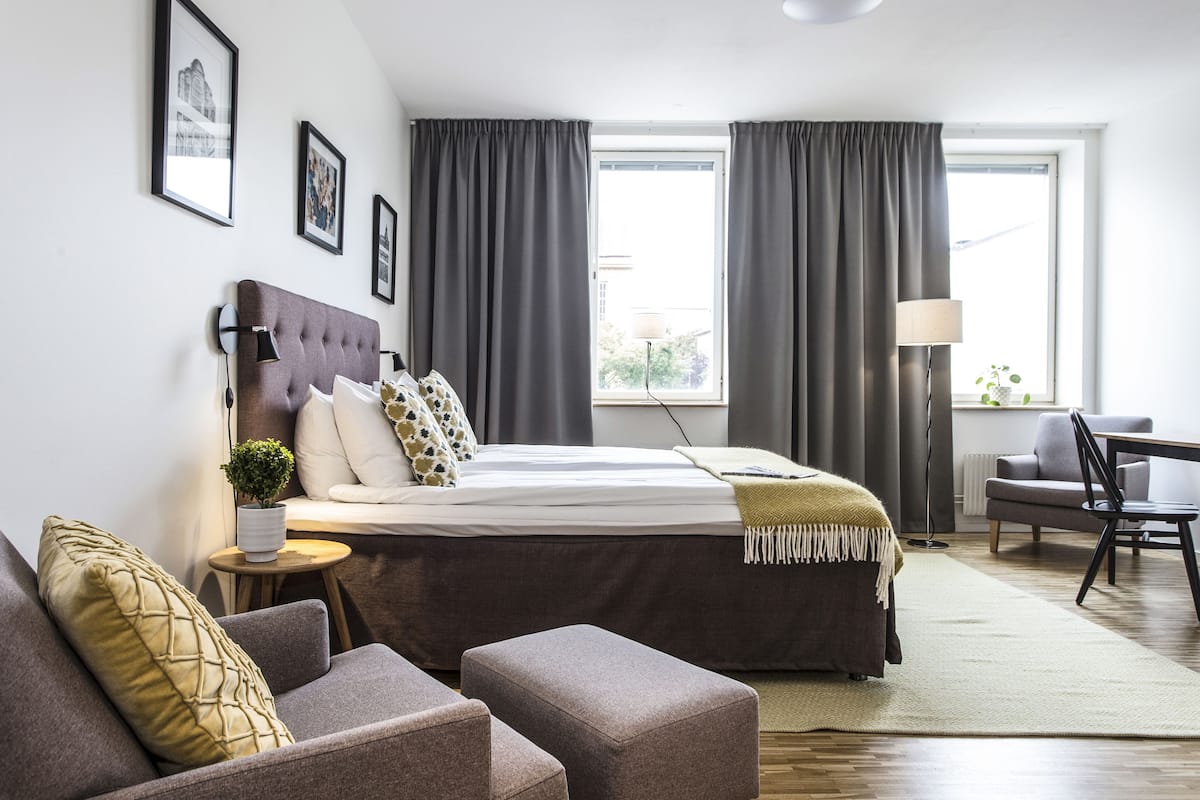 Een moderne hotelkamer met een groot bed, een bank, een kleine tafel en een stoel. Gordijnen hangen voor de ramen, er staat een lamp en er zijn afbeeldingen in lijsten aan de muur.