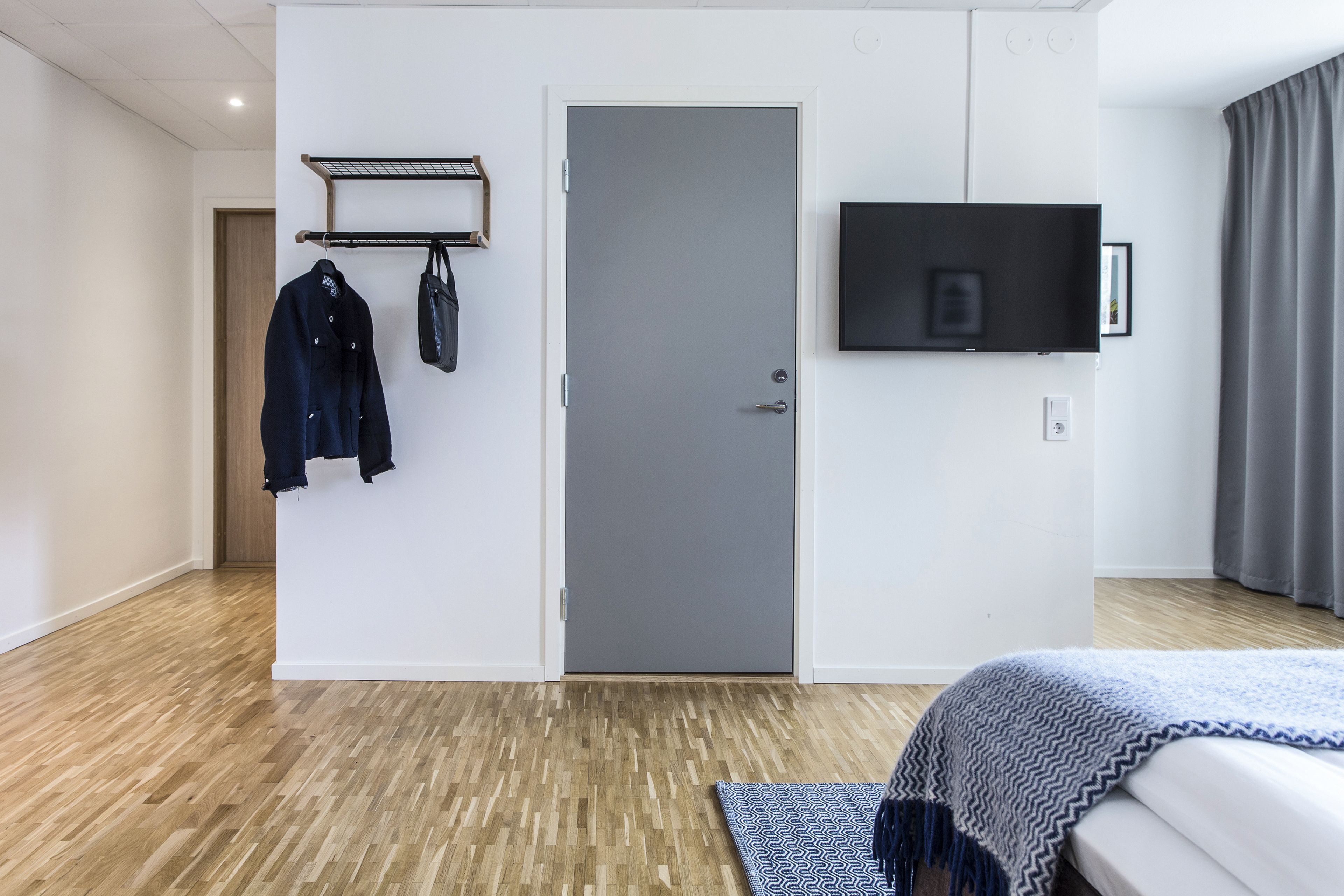 A hotel room with a bed, a grey door, a mounted TV, and a coat rack with jackets.