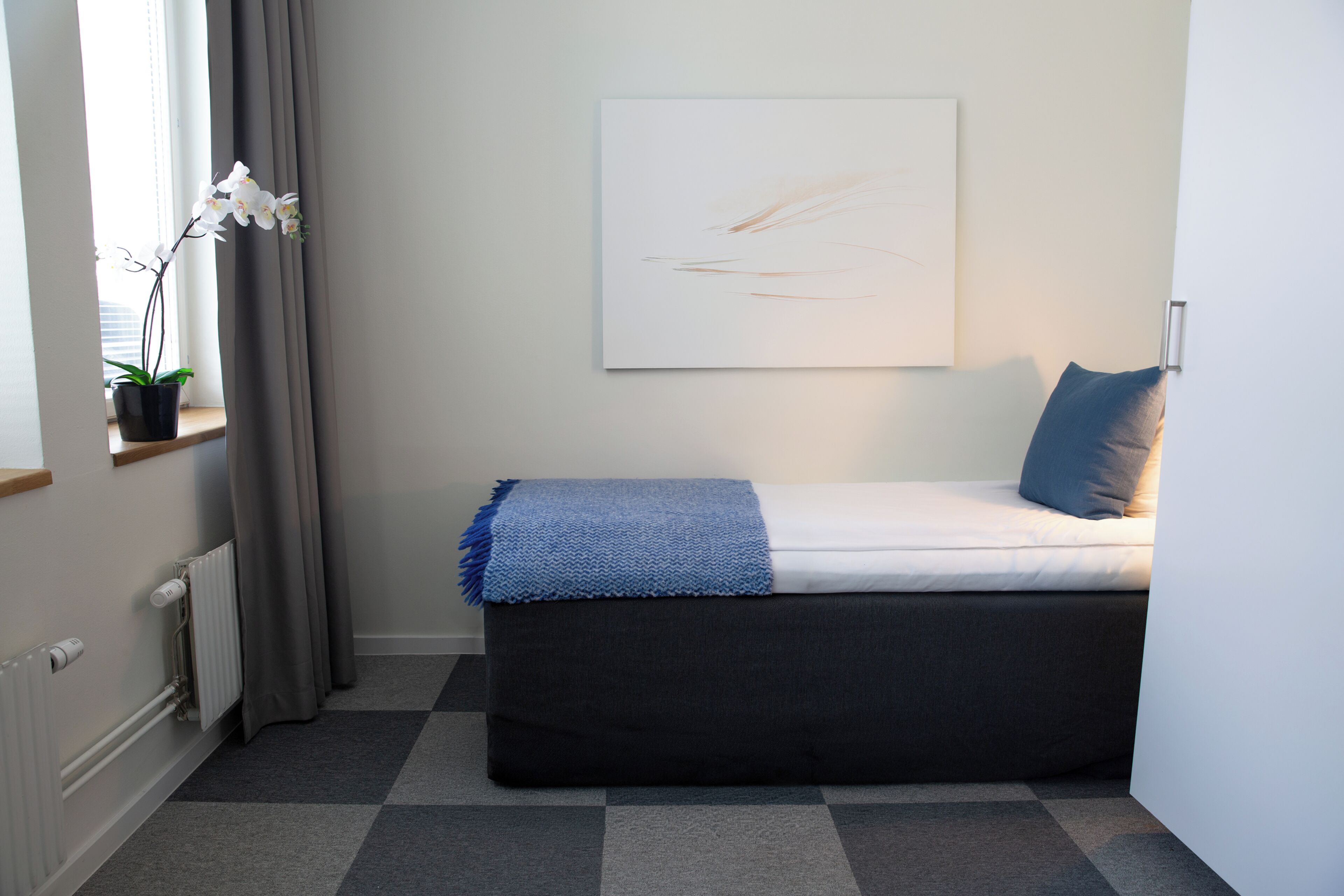 A small, neatly arranged bedroom with a bed, a window with curtains, a radiator, and a wall-mounted artwork.