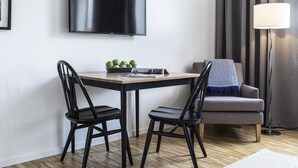 Une chambre d’hôtel moderne équipée d’un téléviseur à écran plat fixé au mur, d’une table à manger avec des chaises, d’un canapé, d’une lampe sur pied et d’un tapis à motifs.