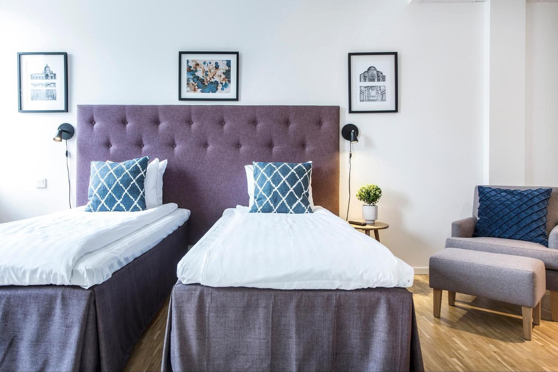 A hotel room with two beds, a grey upholstered headboard, and a grey armchair. There are framed pictures on the wall and a small table with a plant.