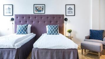A hotel room with two beds, a grey upholstered headboard, and a grey armchair. There are framed pictures on the wall and a small table with a plant.