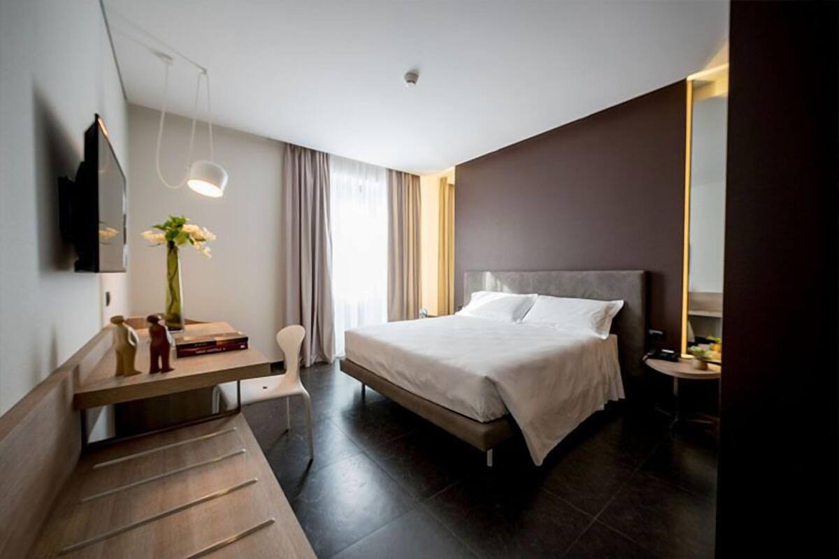 A modern hotel room with a bed, bedside table, chair, and a desk with a lamp.