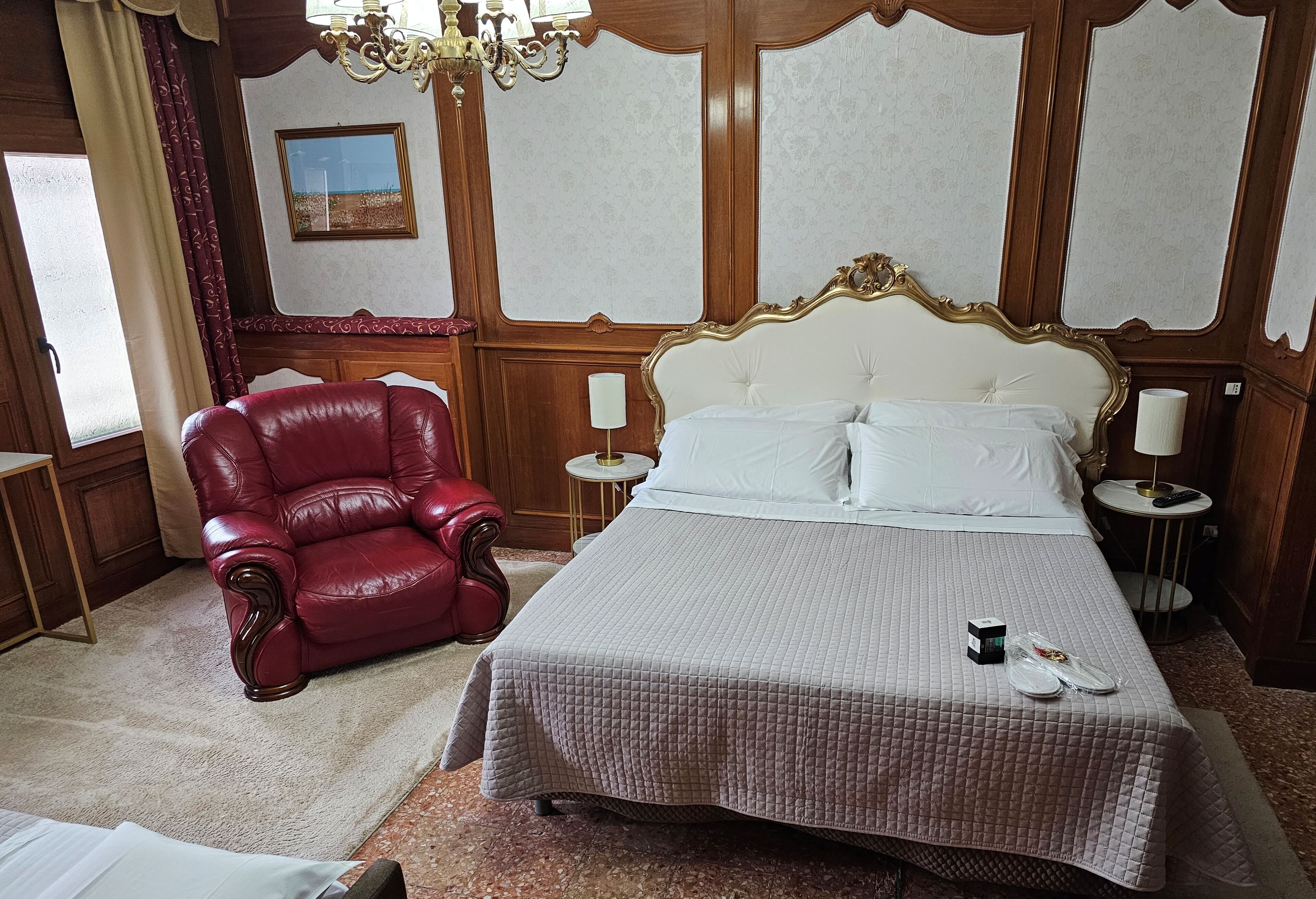 A hotel room with a large bed, a red leather armchair, a chandelier, and a small table with a lamp.