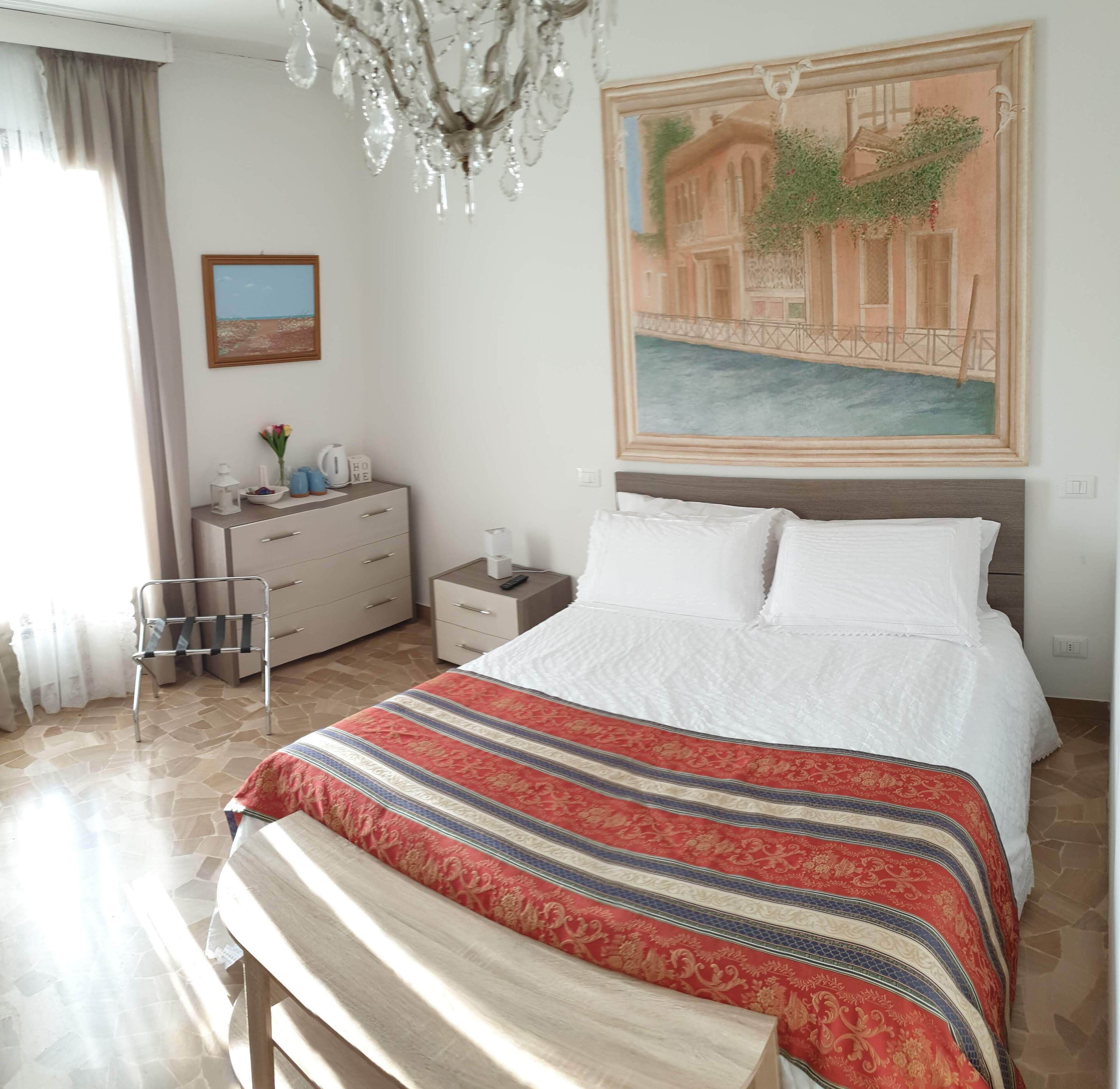 Ein Schlafzimmer mit einem Bett, einer Bank, einem Nachttisch, einer Kommode, einer Chandelierleuchtre und einem Wandbild.