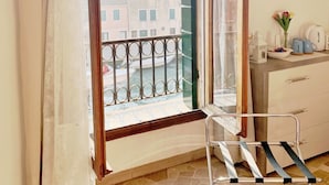 A room with a balcony, a window with curtains, a small table with a vase and teapots, and a chair.