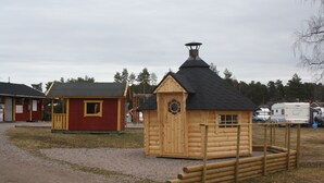 A small wooden cabin with a black roof and a chimney, surrounded by a wooden fence. - Årsunda Strandbad (Arsunda)