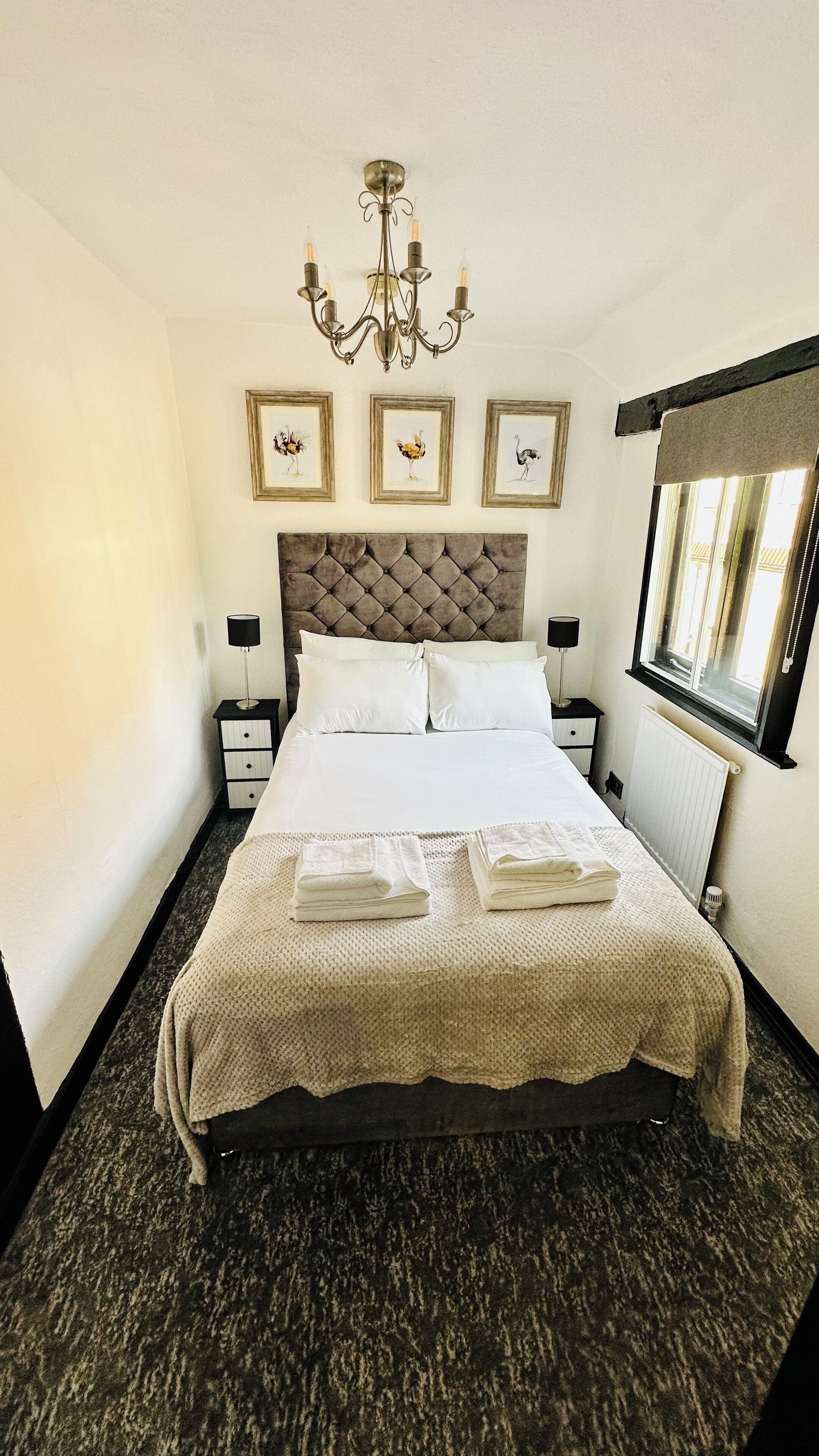 A neatly made bed with a tufted headboard, a chandelier, framed artwork on the wall, and a window with a view.
