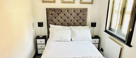 A neatly made bed with a tufted headboard, a chandelier, framed artwork on the wall, and a window with a view.